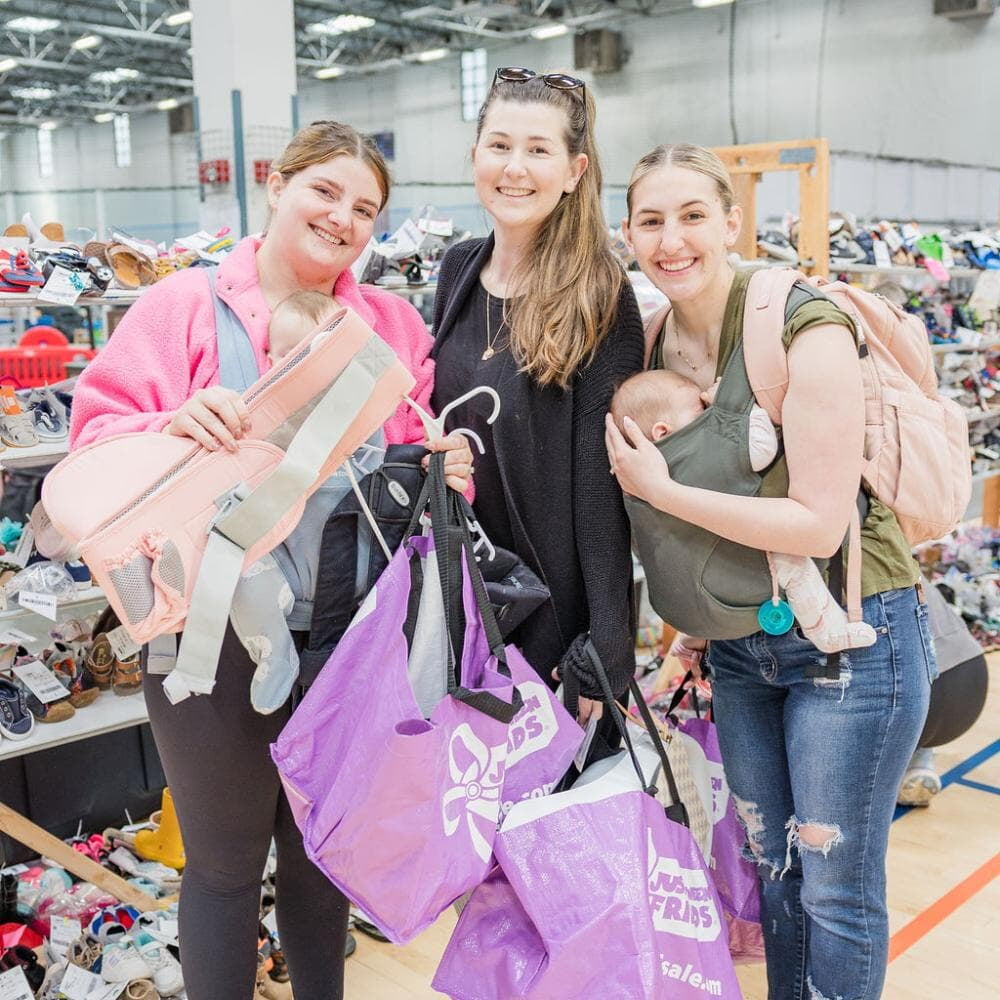 Three ladies shopping with purple JBF bags filled with items. Two of the women have babies with them.