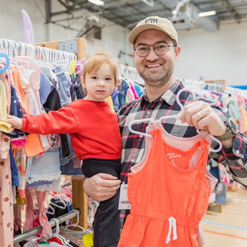 A father holds his daughter in one arm and is holding hangers of clothing in his other hand.