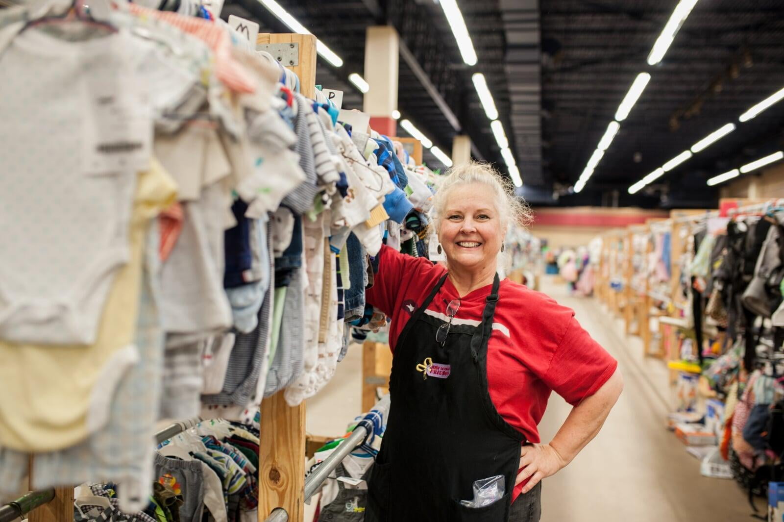 A JBF Team Member wears a black JBF apron while organizing infant clothing on the clothing racks.