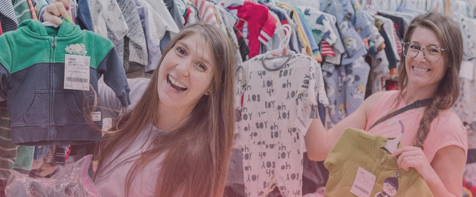 Two ladies smile while holding up baby clothing while shopping.