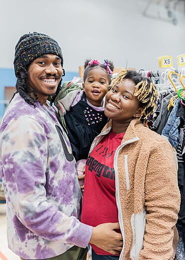 Dad has a kit hat on and a white and purple tie-dye sweatshirt. Mom has a red shirt and tan jacket and holds a toddler girl.